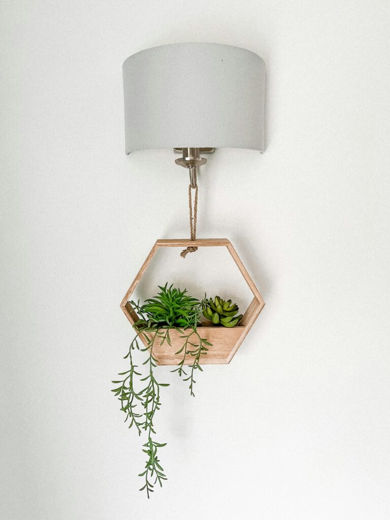 Modern hexagonal wooden shelf with succulents and a lamp on a white wall.